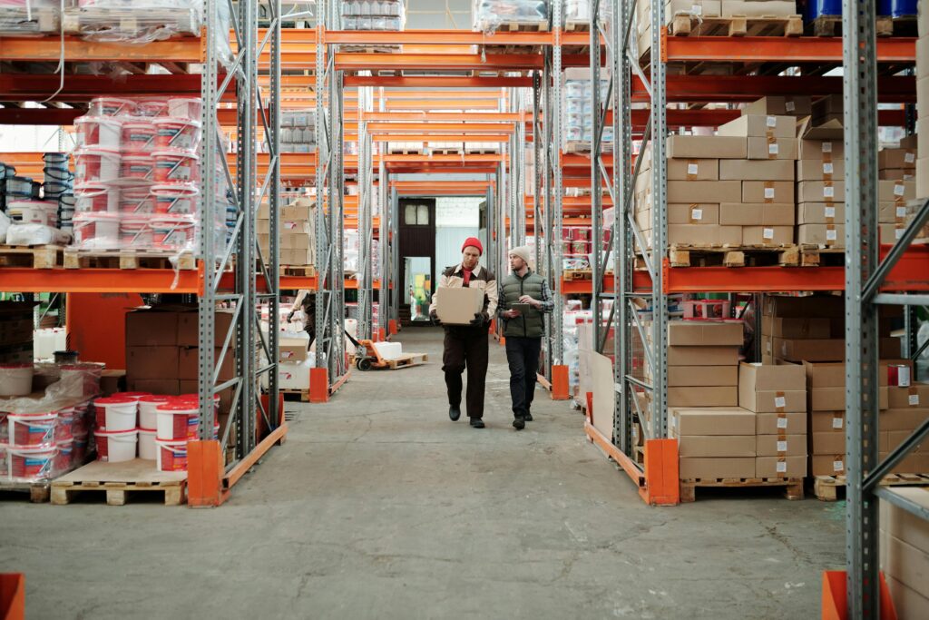 Two workers handle a package in a spacious warehouse surrounded by shelves stocked with boxes and products.